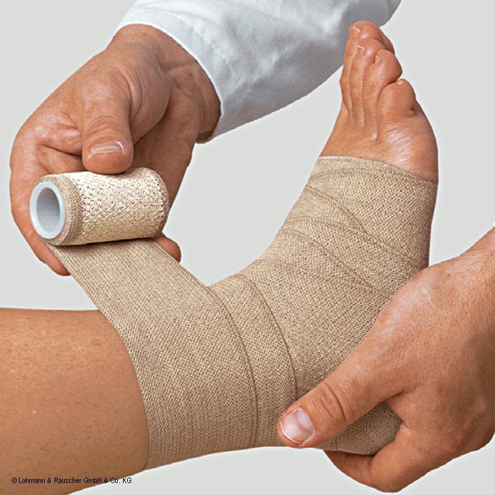 Beige elastische Bandage wird um einen Fuß gewickelt. Hände halten Rolle und wickeln Bandage. Fuß und Unterschenkel sind sichtbar.