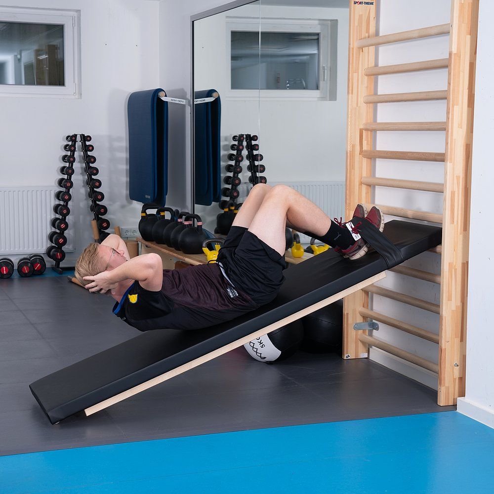 Schrägbrett mit Person. Schwarze gepolsterte Oberfläche, Holzrahmen. Person liegt auf dem Brett, trainiert. An der Wand ist eine Sprossenwand.