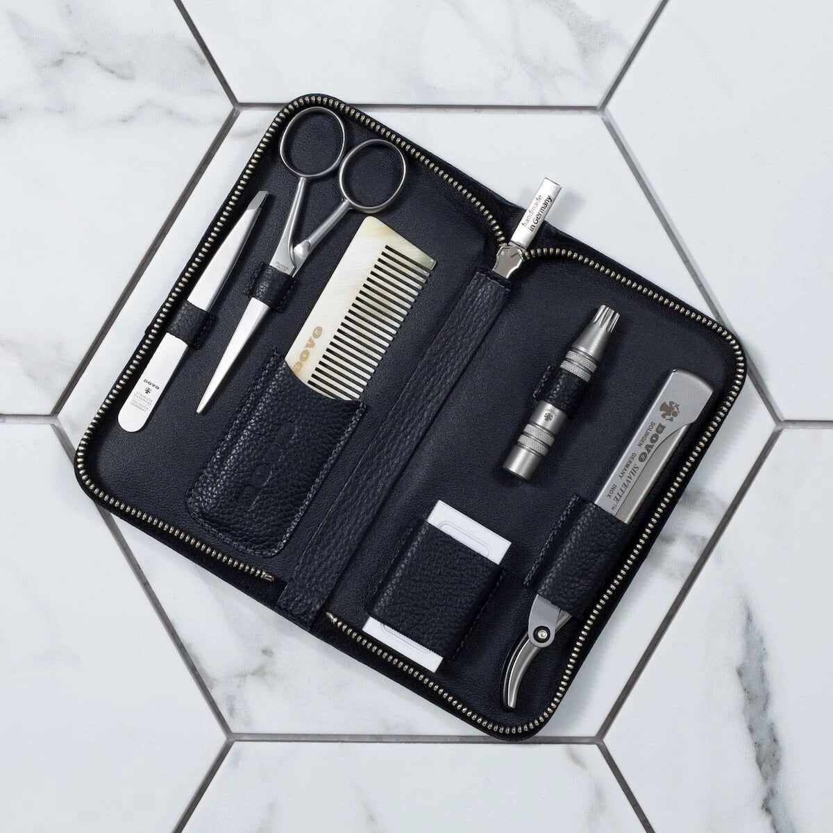 Schwarzes Etui mit Reißverschluss, gefüllt mit Bartpflege-Werkzeugen: Schere, Kamm, Pinzette, Nasenhaartrimmer, Nagelknipser und Etui. Auf Fliesen.