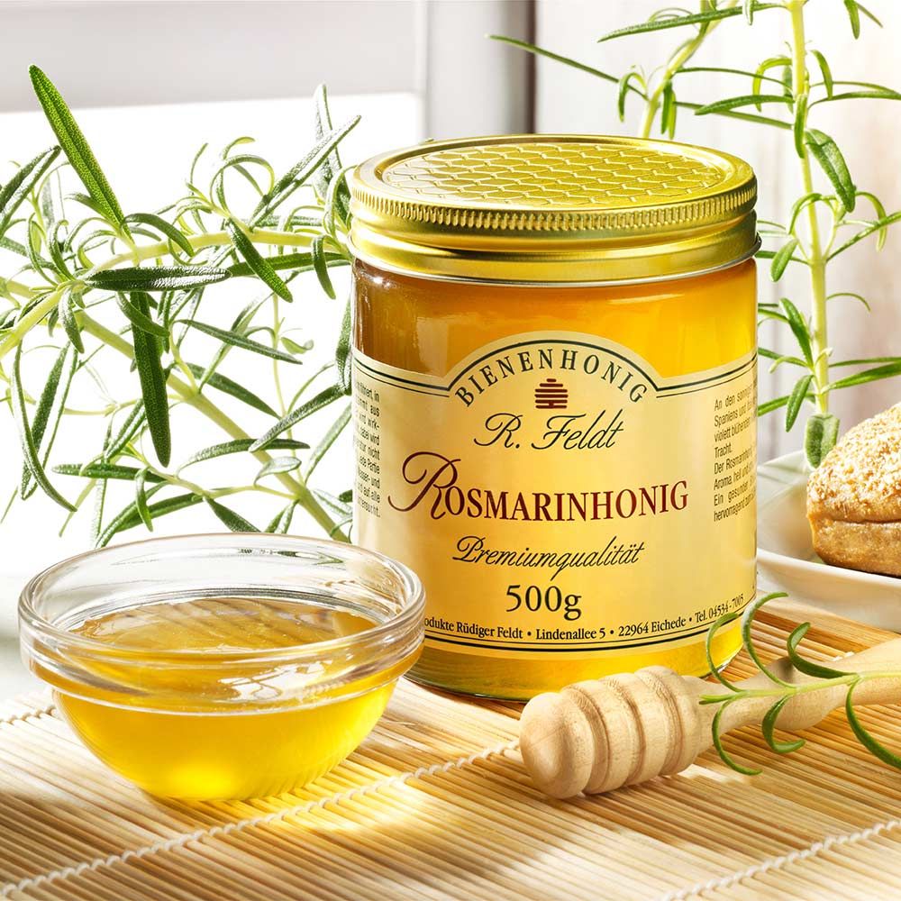 Glas Honig mit Etikett "Rosmarinhonig" und "500g". Goldfarbener Deckel. Honigschale und Honiglöffel daneben.