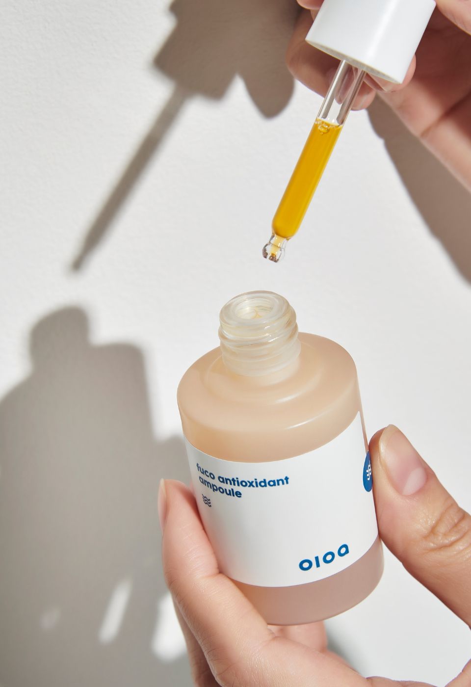 Kleine, rosa Flasche mit Pipette, die eine gelbe Flüssigkeit enthält. Flasche mit Aufkleber und Logo. Marke: OIOA. Hände halten die Flasche.