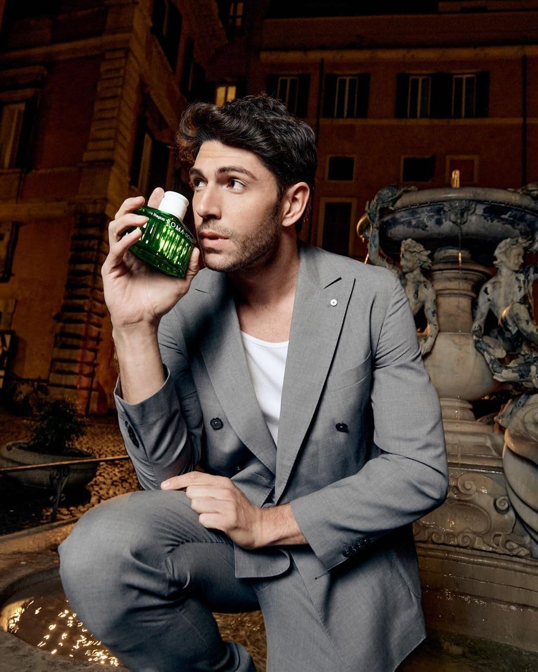 Mann hält grüne Flasche. Im Hintergrund Brunnen und Gebäude. Flasche mit Aufschrift: ROMA UOMO.