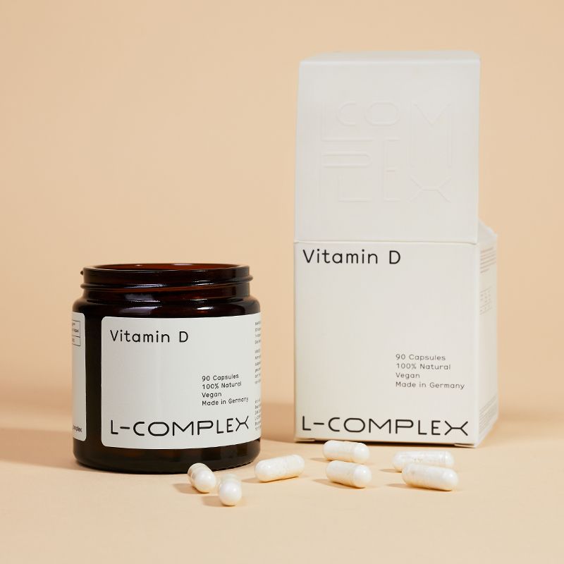 Braune Glasdose und Schachtel mit "Vitamin D" und "L-COMPLEX". 90 Kapseln, vegan. Einige Kapseln liegen daneben.