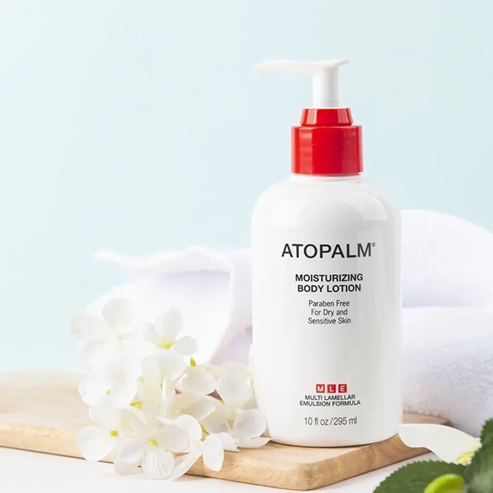 Weiße Flasche mit rotem Pumpkopf, neben Blumen und Handtuch. Aufschrift: ATOPALM Moisturizing Body Lotion.