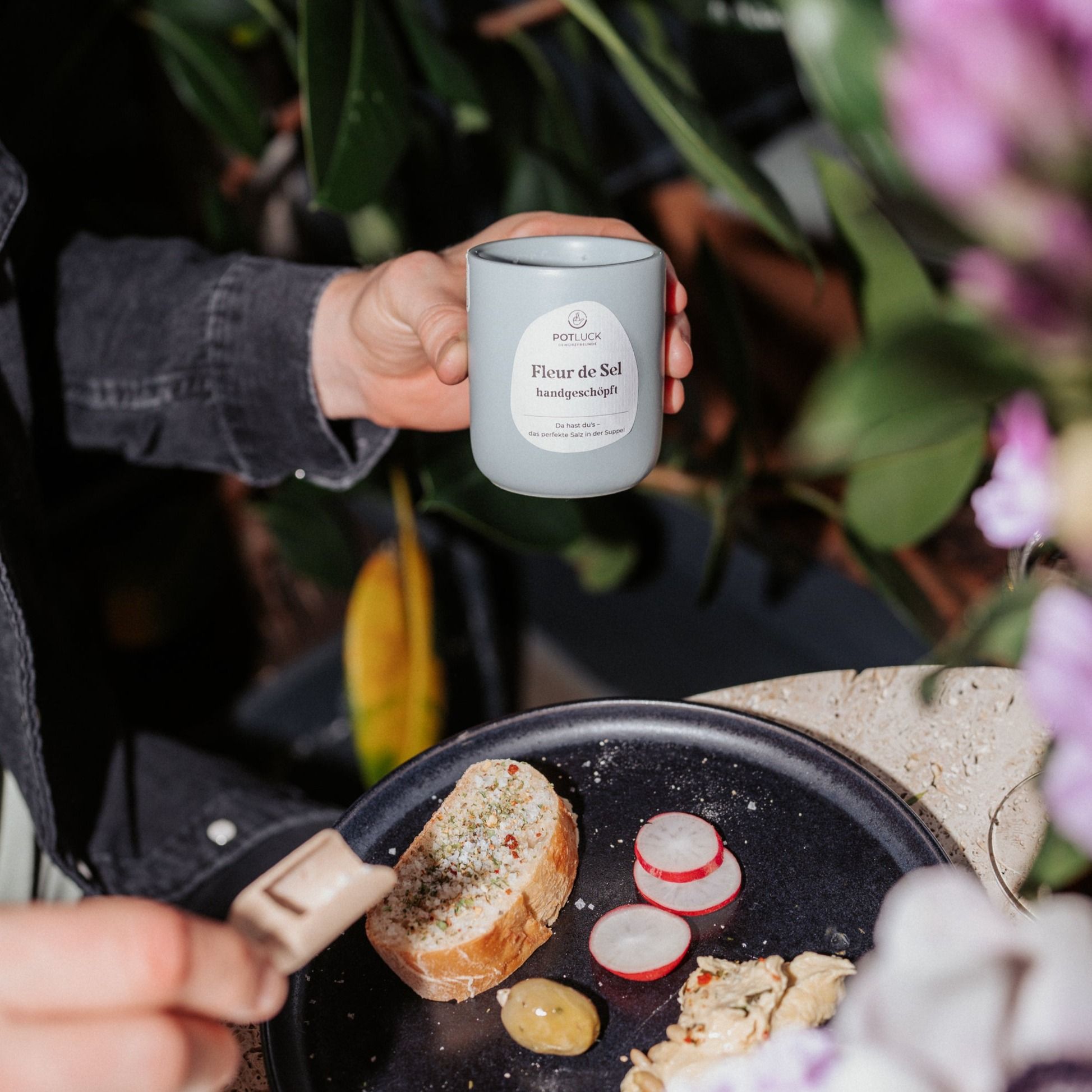 Person hält hellblaue Dose mit "POTLUCK Fleur de Sel handgeschöpft"-Etikett. Essen auf Teller.