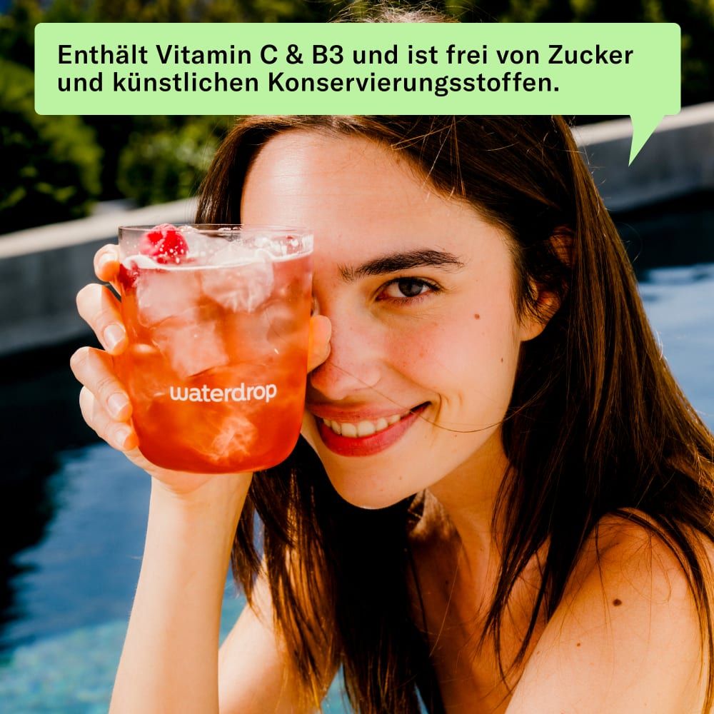 Frau hält Glas Himbeer-Eistee vor dem Gesicht. Auf dem Glas: waterdrop®. Im Hintergrund: Pool.