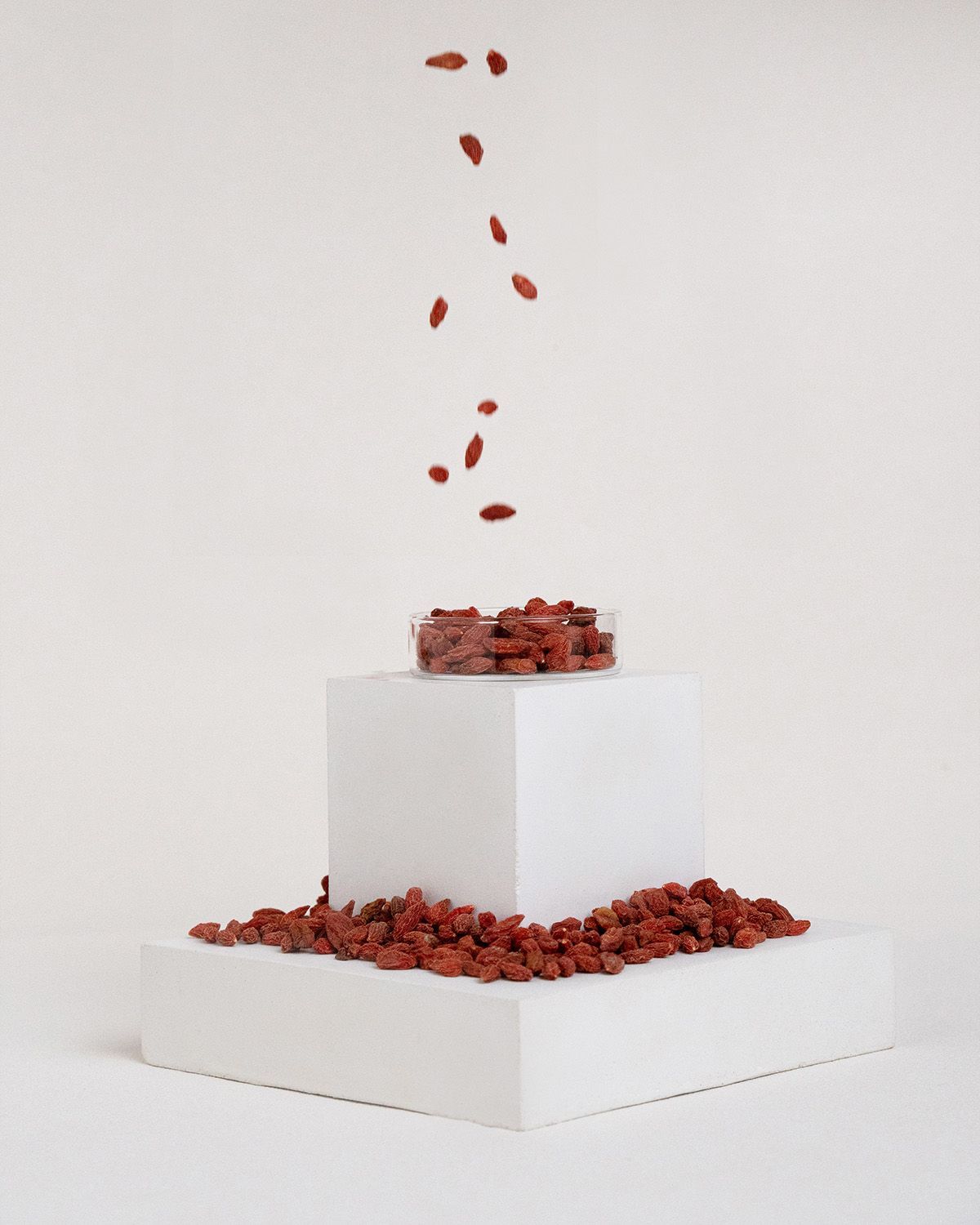 Getrocknete Goji-Beeren in einer transparenten Schale auf einer weißen Plattform. Einige Beeren fallen herunter.