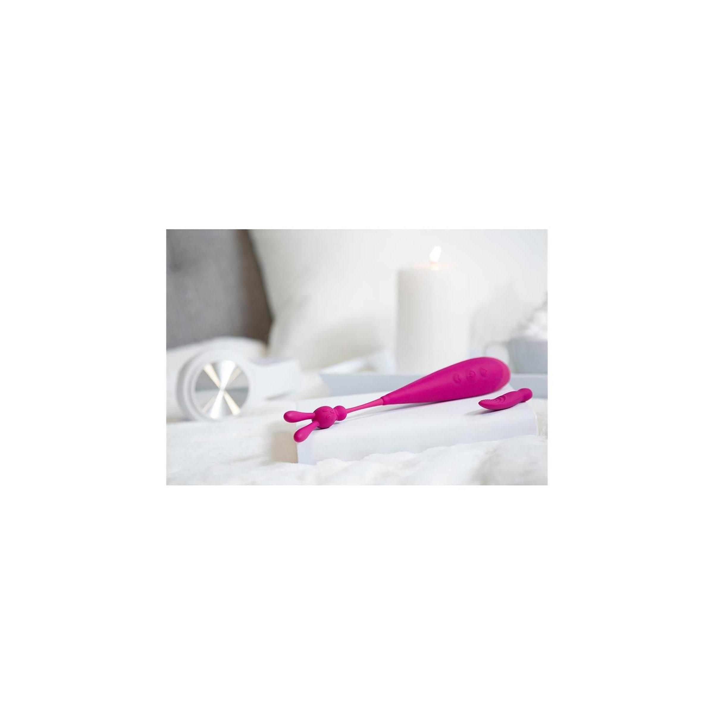 Rosa Vibrator mit zwei Aufsätzen auf weißer Oberfläche. Kerze und Kopfhörer im Hintergrund.