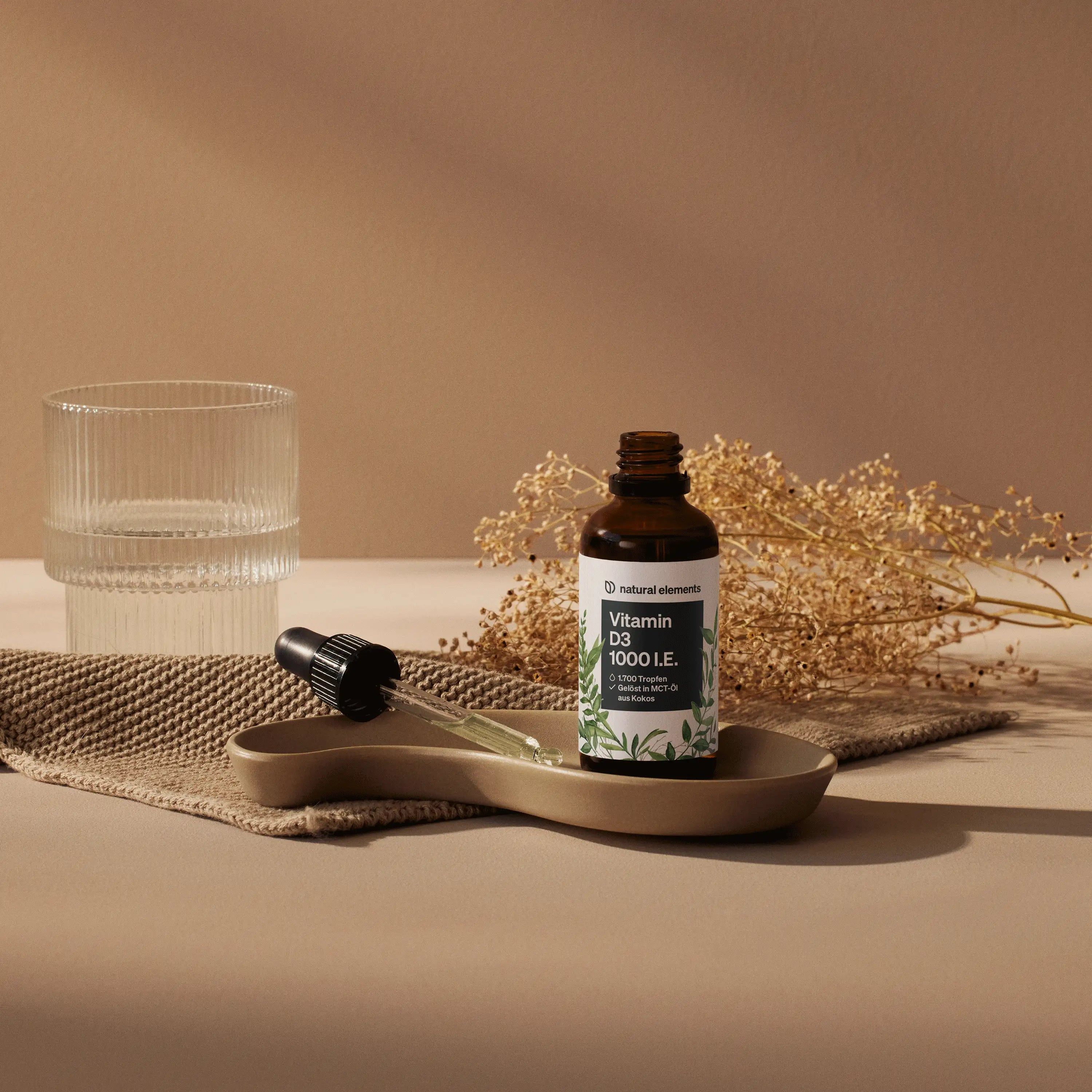 Braune Flasche mit "Vitamin D3 1000 I.E." in einer Schale mit Pipette. Glas Wasser daneben.