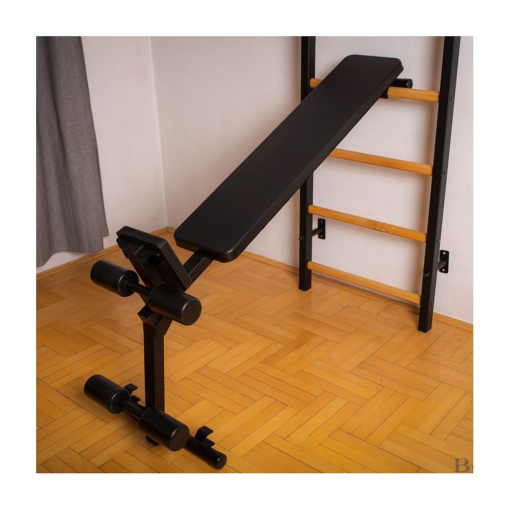 Schwarze Trainingsbank vor Sprossenwand. Verstellbare Rückenlehne und Beinpolster. Metallrahmen. Holzfußboden im Hintergrund.