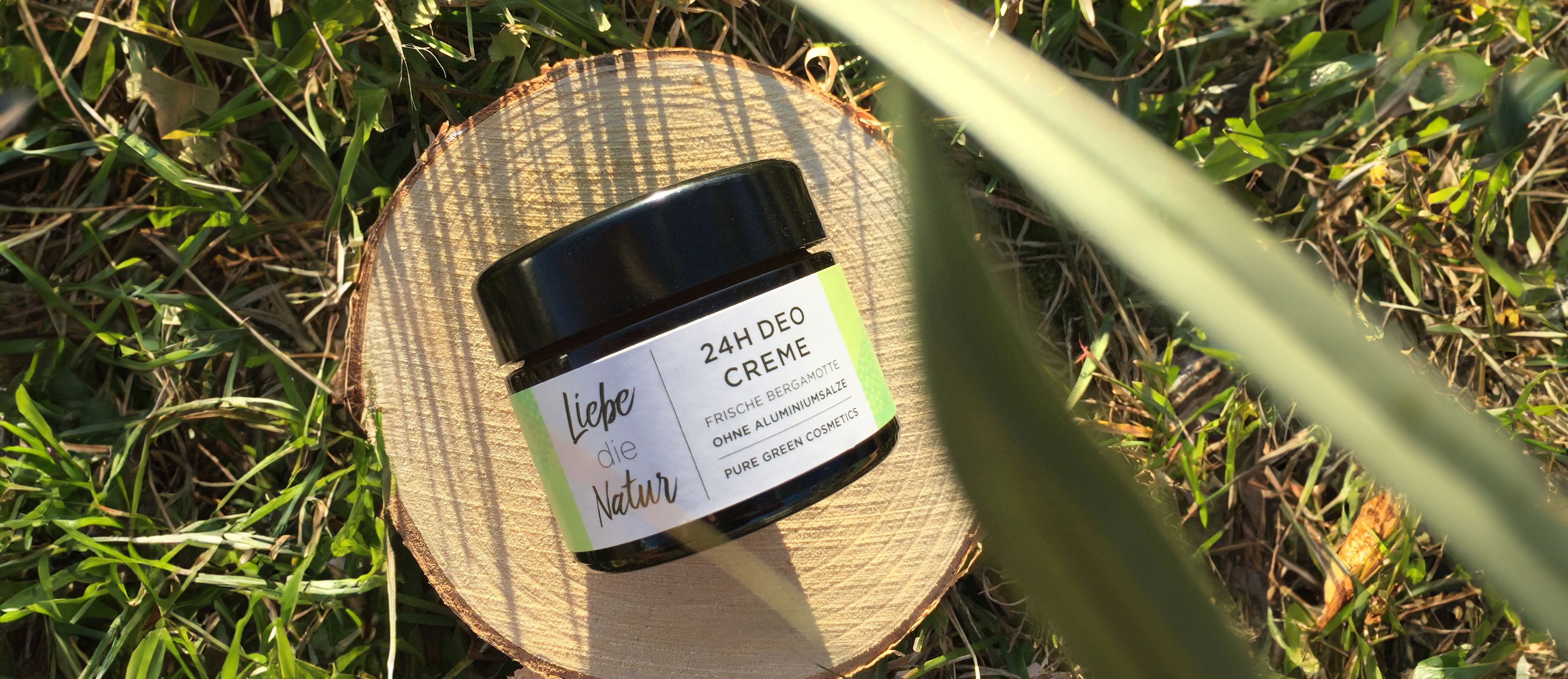 Deocreme-Glas mit schwarzem Deckel auf Holzscheibe. Etikett mit Schriftzug und grünen Akzenten. Natürliche Umgebung mit Gras.