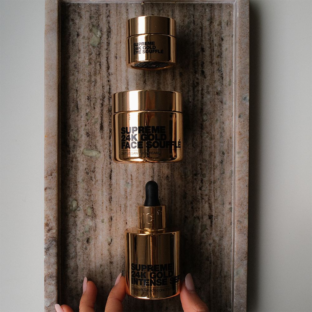 Drei goldene Behälter auf einem Tablett. Produktnamen: SUPREME 24K GOLD FACE SOUFFLÉ, SUPREME 24K GOLD INTENSE SERUM, SUPREME 24K GOLD EYE SOUFFLÉ.