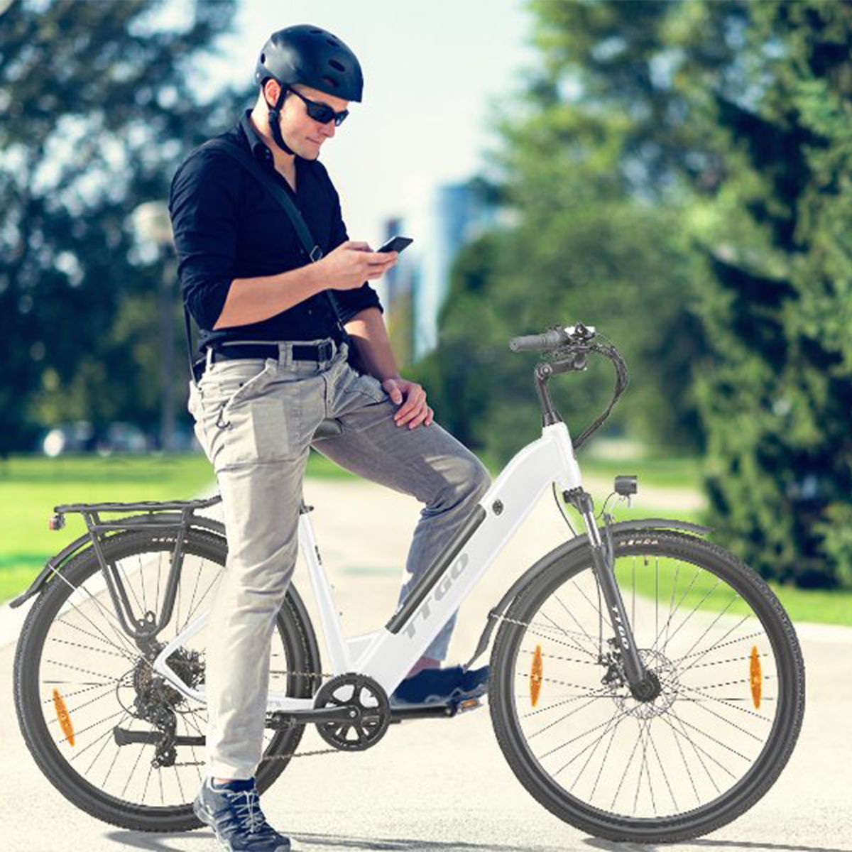 Mann steht neben weißem E-Bike. Helm, Sonnenbrille und Smartphone. Hintergrund: Bäume und Gebäude.
