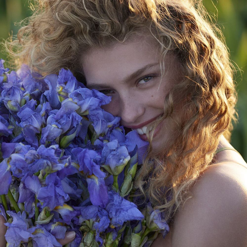 Porträt einer Frau mit lockigem Haar, die einen Strauß blauer Blumen umarmt. Sie lächelt in die Kamera.