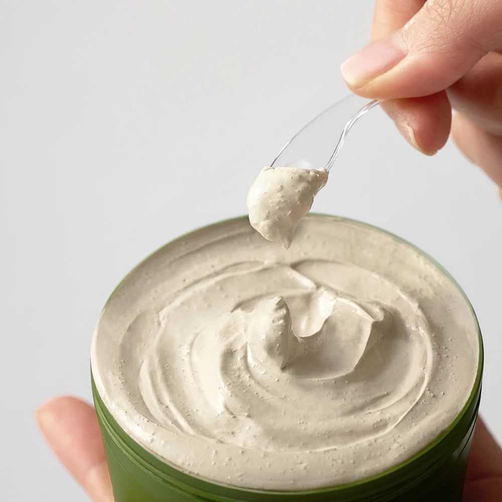 Grüne Dose mit weißer Gesichtsmaske. Eine Hand hält einen Spatel mit Maske. Der Hintergrund ist weiß.