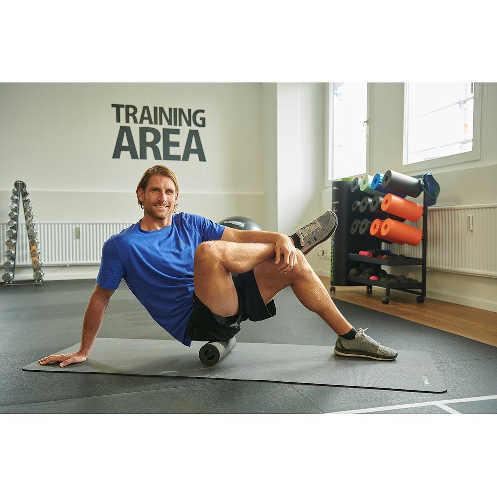 Mann trainiert im Fitnessstudio. Er sitzt auf einer Matte und rollt mit einer Faszienrolle. Im Hintergrund: Trainingsbereich.