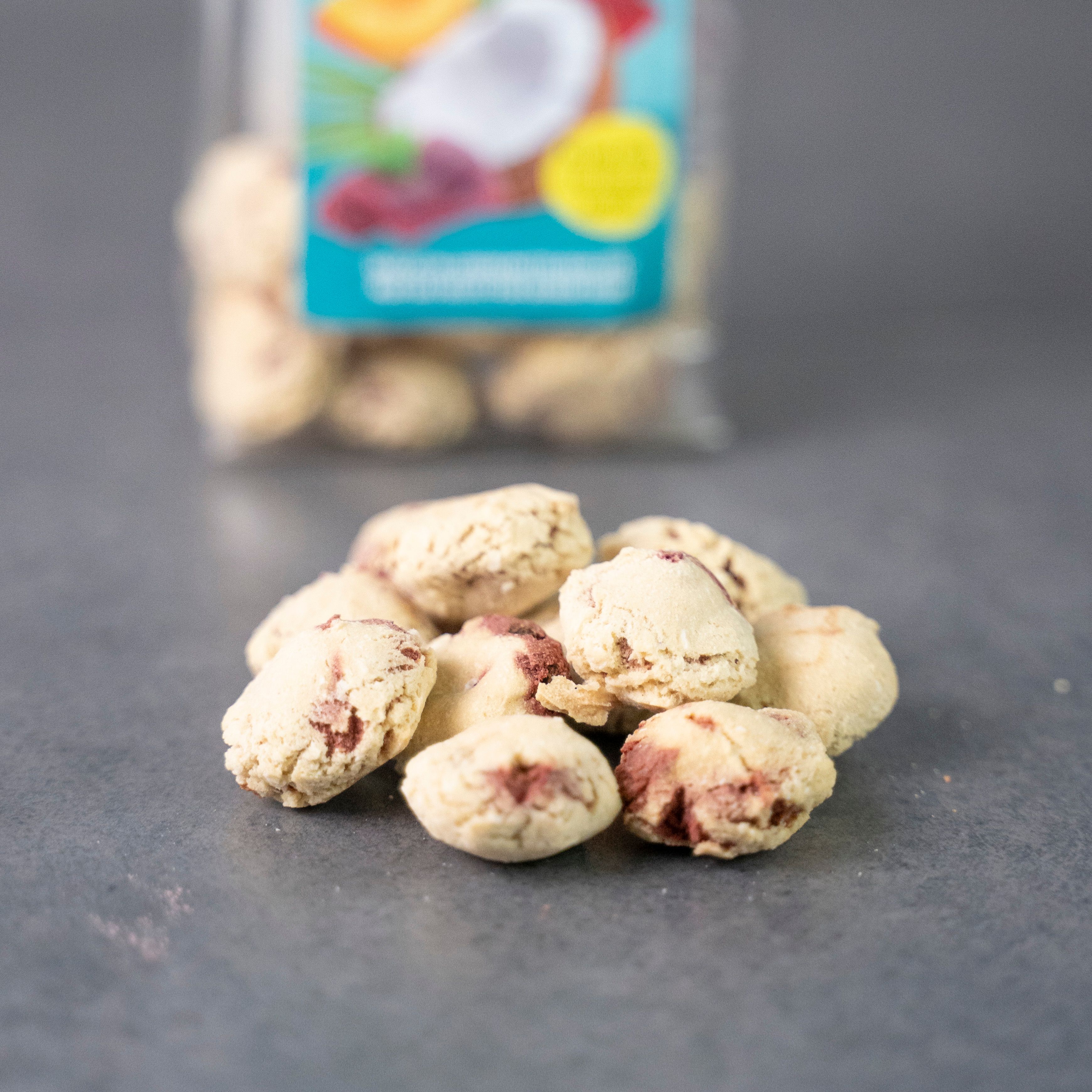 Haufen Kokos Crunchies auf grauem Untergrund. Die Crunchies sind hellbraun mit roten Flecken. Im Hintergrund eine transparente Tüte.