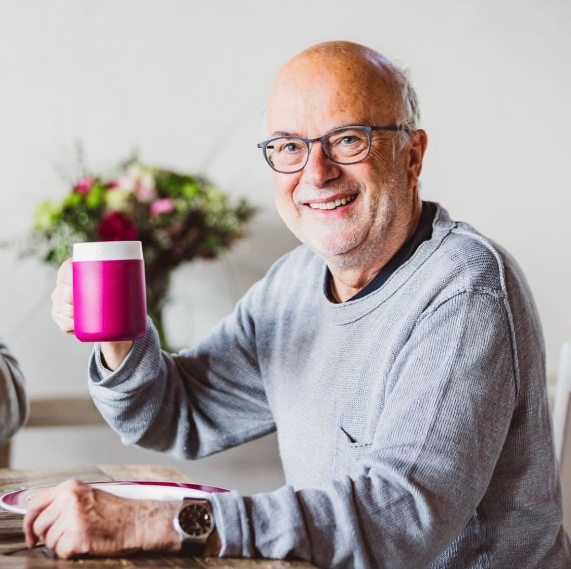 Mann hält einen pinkfarbenen Becher mit großem Henkel. Er sitzt an einem Tisch und lächelt. Im Hintergrund Blumen.