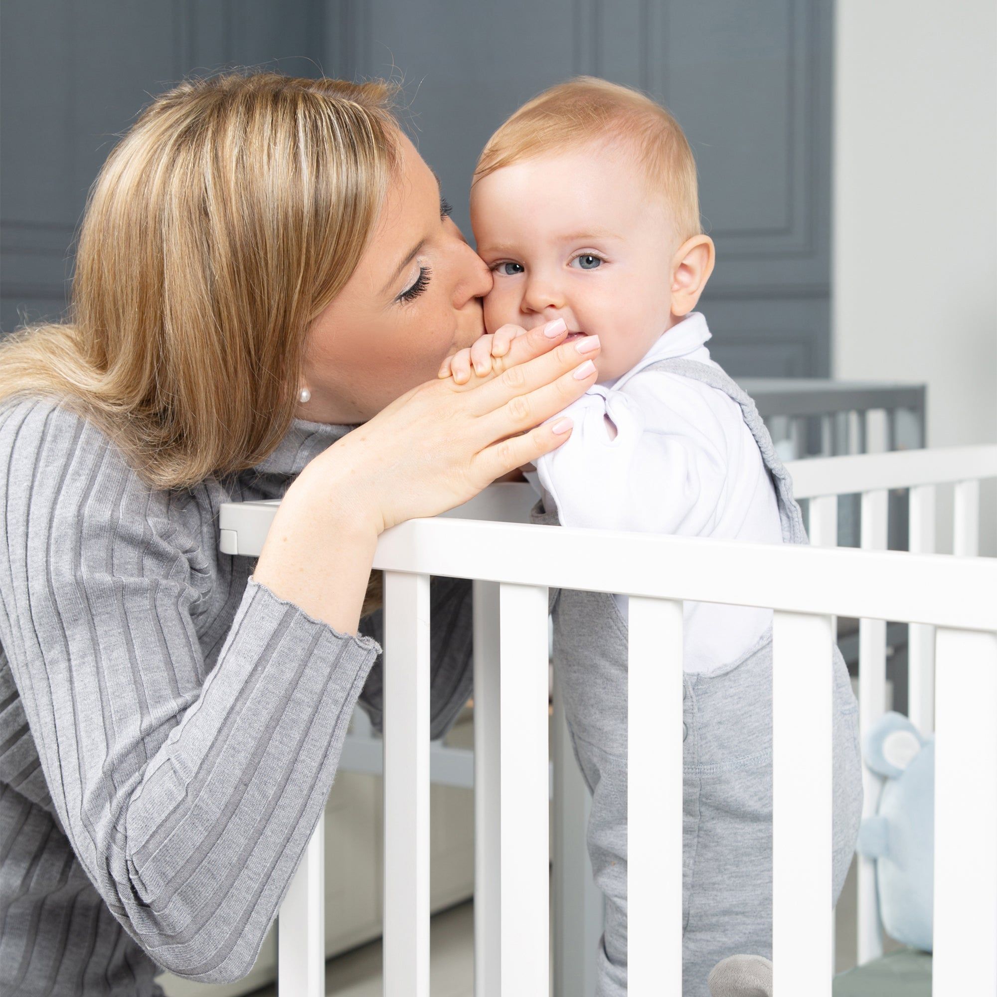 Mutter küsst Baby in weißem Laufgitter. Baby trägt graue Kleidung. Grauer Hintergrund.