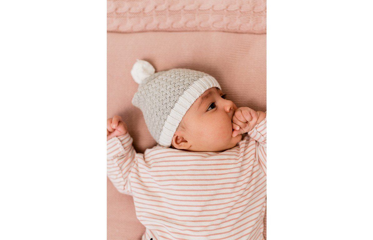 Baby mit grau-weißer Bommelmütze. Strickmuster, Bommel oben. Baby liegt auf rosa Untergrund.