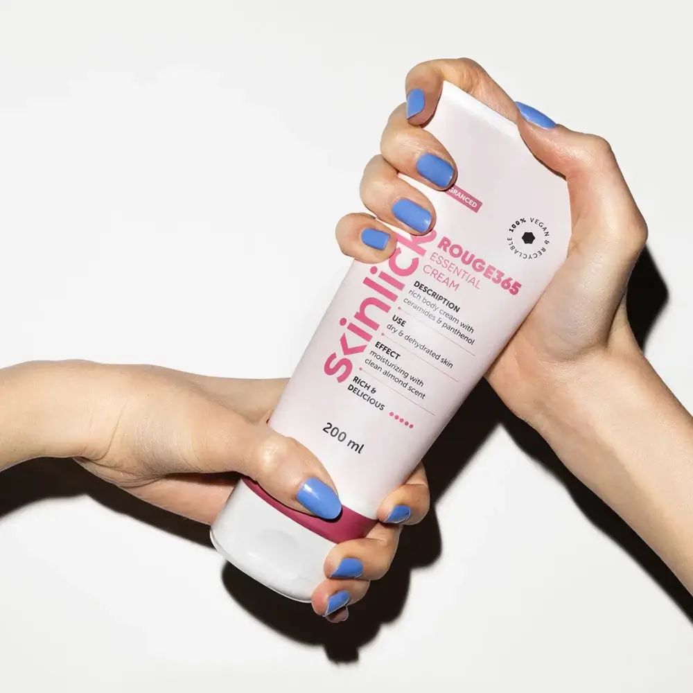 Hände halten eine Tube SKINLICK Rouge365 Essential Cream. Rosa Tube mit weißem Deckel. Blaue Fingernägel.