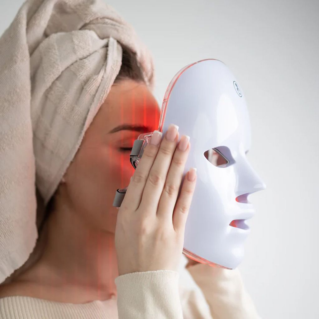 Eine Frau hält eine weiße LED-Gesichtsmaske an ihr Gesicht. Die Maske hat Öffnungen für Augen, Nase und Mund. Rotes Licht.