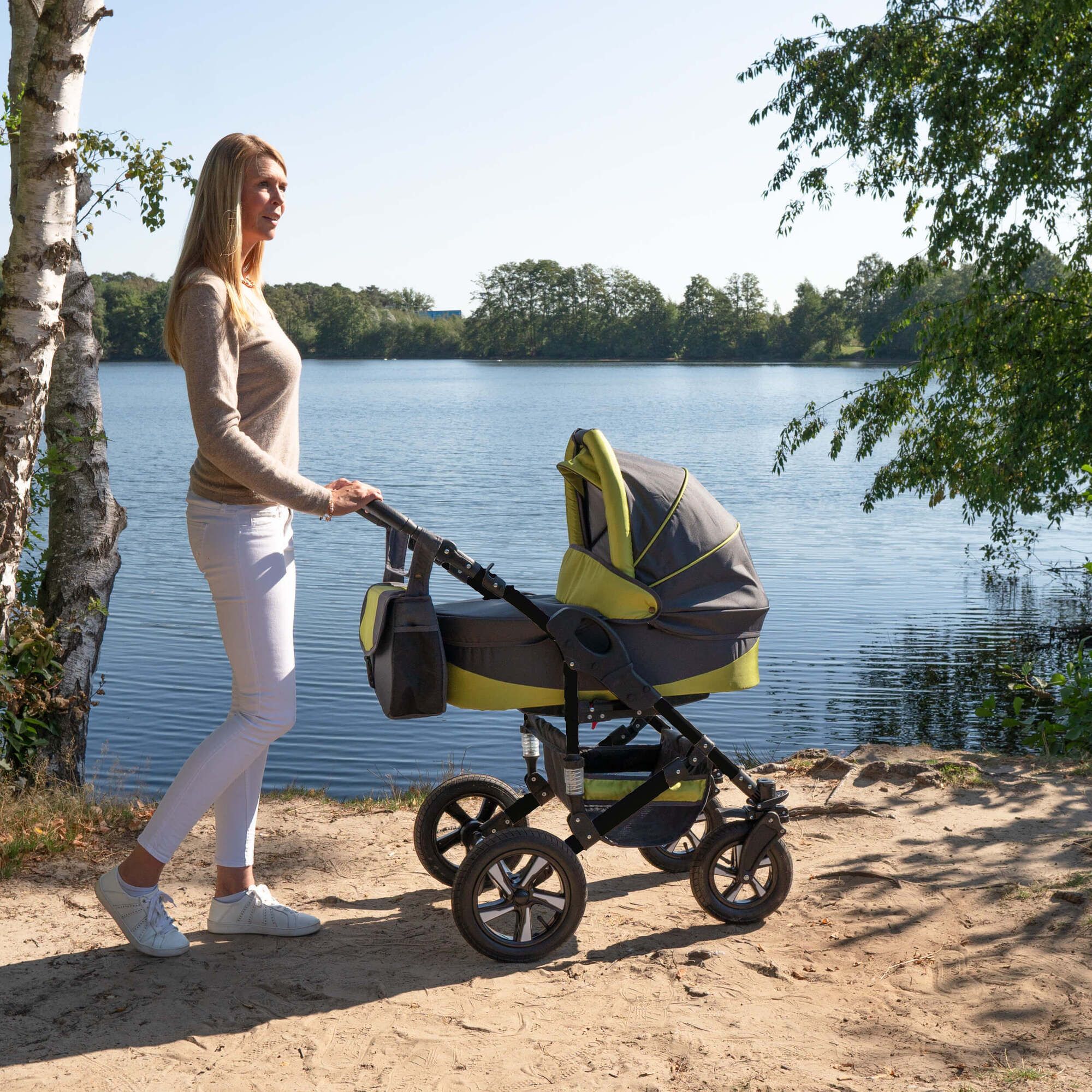 Frau schiebt Kinderwagen, grau-grün, am Seeufer. Schwarzes Gestell, Hartgummireifen. Tasche am Griff. Sonniger Tag.