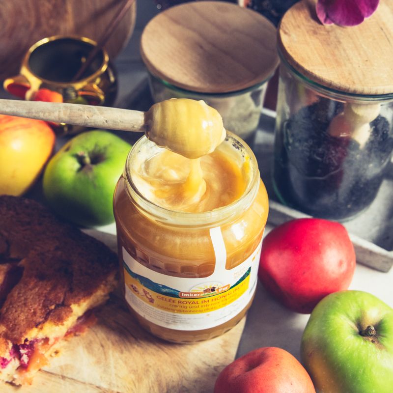 Glas Honig mit Etikett, Löffel mit Honig. Aufschrift: Gelee Royal im Honig (roh). Goldener Deckel, weißes Etikett. Neben Äpfeln und Kuchen.