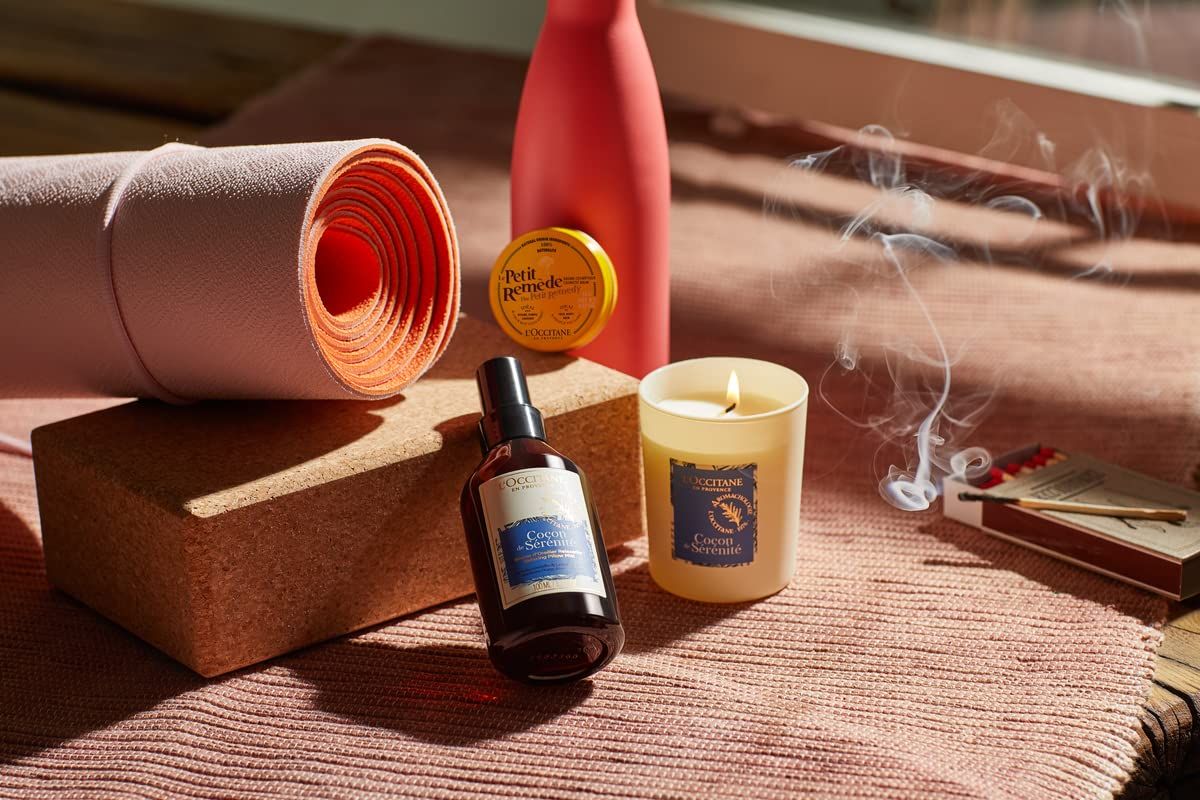 Braune Flasche mit schwarzem Sprühkopf neben Kerze, Yogamatte und Block. Etikett mit Produktnamen. L'Occitane Aromachologie Kopfkissen-Nebel Entspannend.