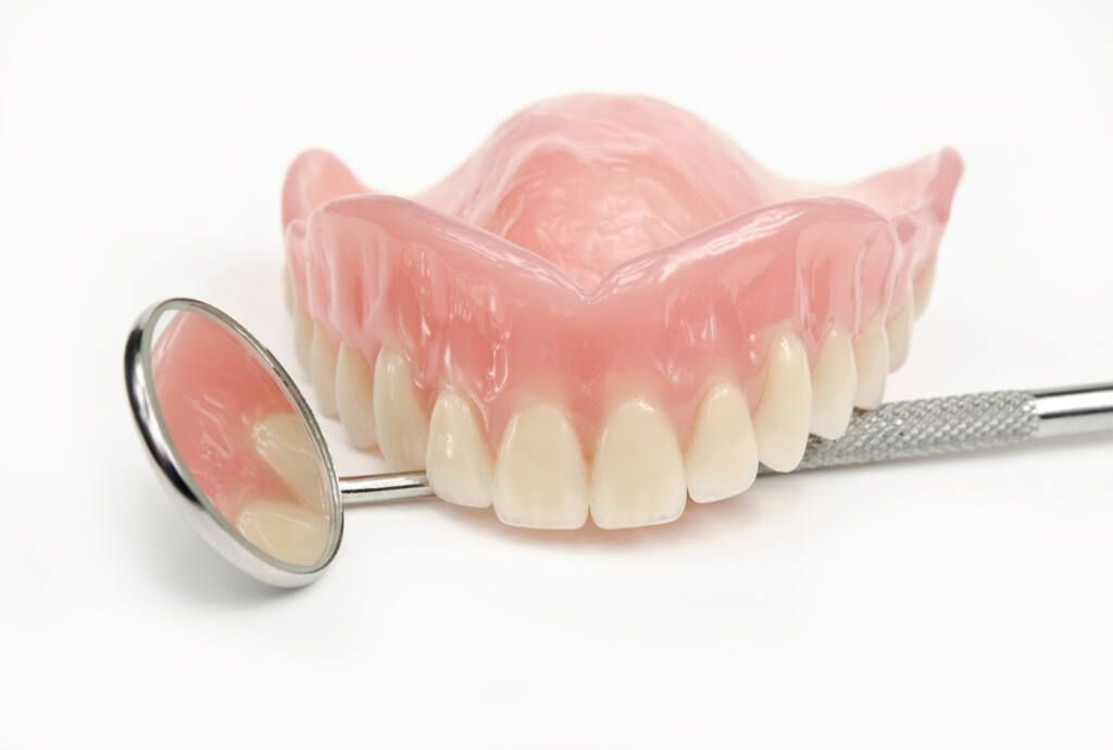 Zahnprothese mit Zahnarztspiegel. Prothese mit rosa Basis und weißen Zähnen. Spiegel reflektiert Zähne.