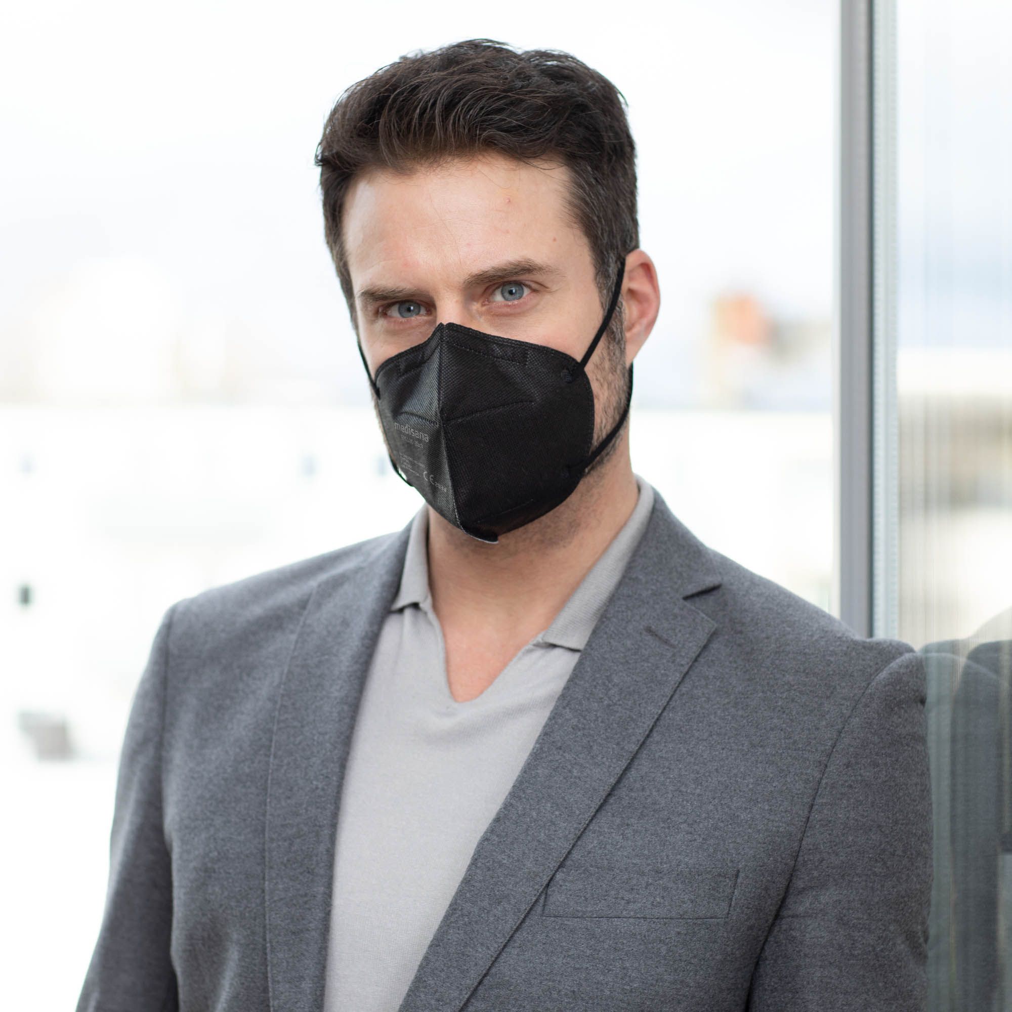 Mann trägt schwarze FFP2-Atemschutzmaske, grauer Blazer, graues Hemd, Fenster im Hintergrund.