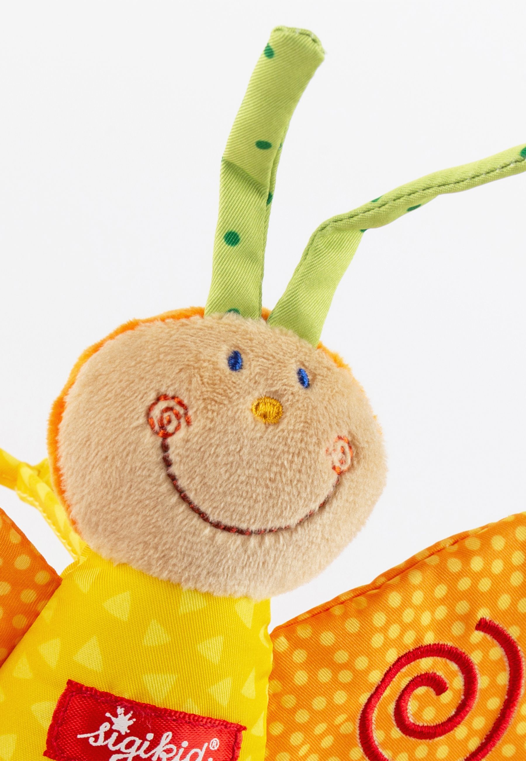 Nahaufnahme des Gesichts eines Stoff-Schmetterlings. Orangefarbener Kopf, grüne Fühler. Rotes Label mit Logo.