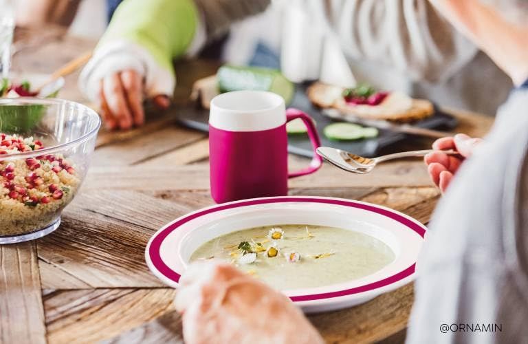 Teller mit rotem Rand, gefüllt mit Suppe. Person hält Löffel. @ORNAMIN am unteren Rand.