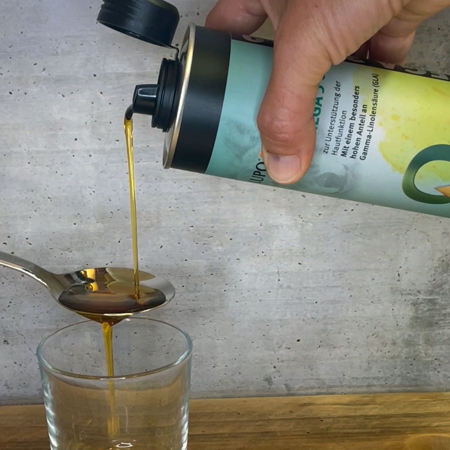 Hand gießt Öl aus einer Dose in einen Löffel und ein Glas. Dose mit Aufschrift: OMEGA 3-6-9. Öl ist goldfarben.