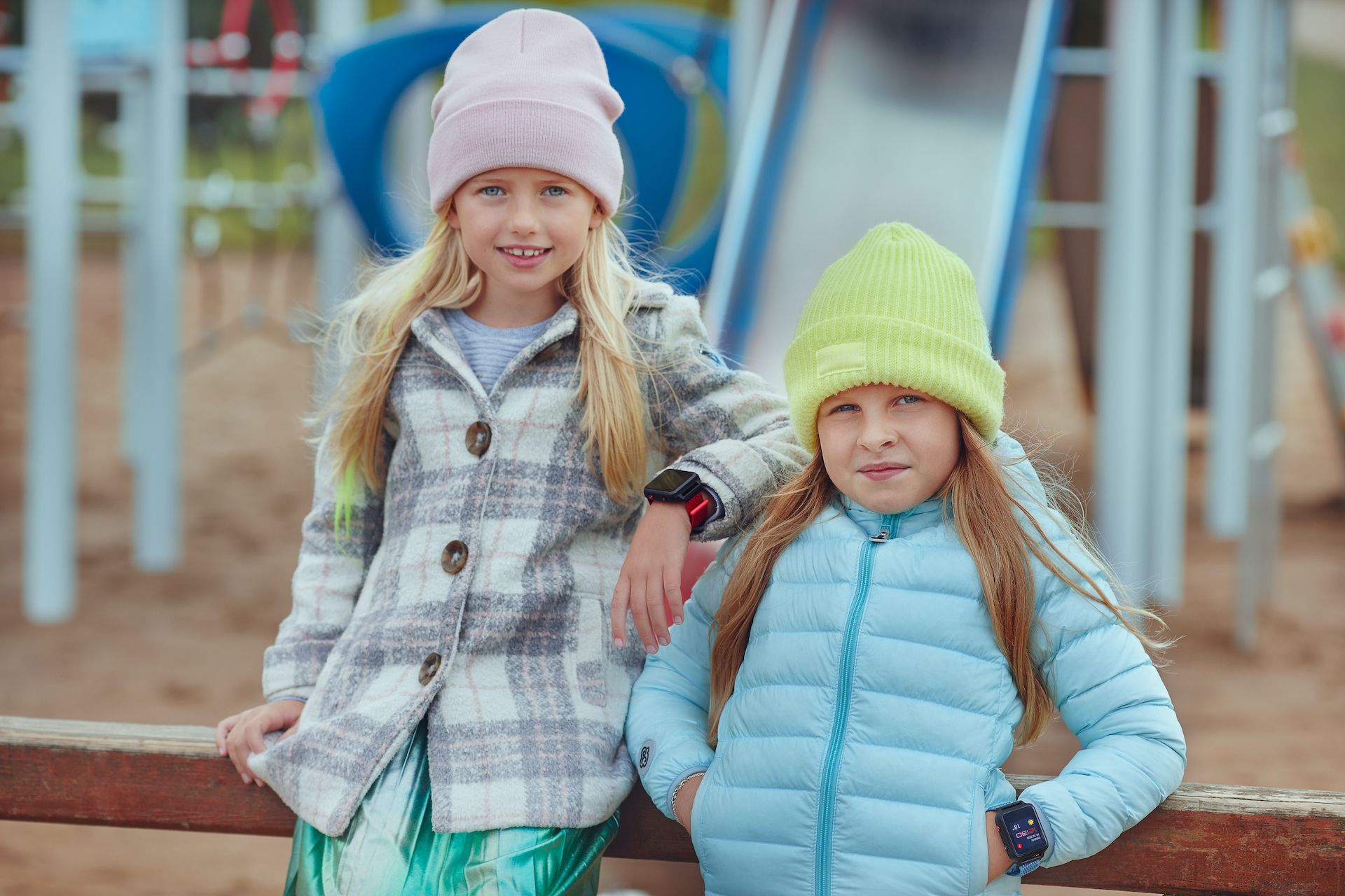Zwei Mädchen mit Mützen und Jacken. Beide tragen Smartwatches am Handgelenk. Hintergrund: Spielplatz.