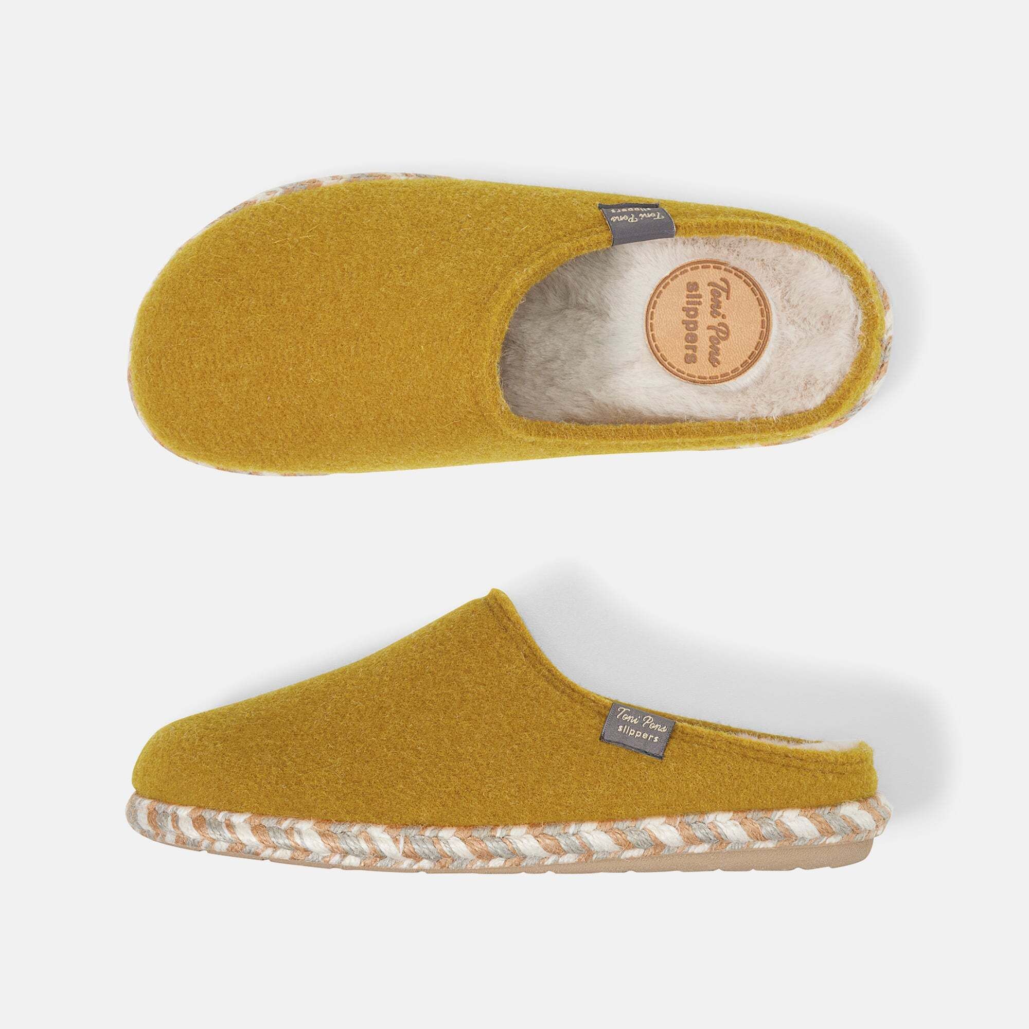 Zwei senffarbene Hausschuhe mit weißem Innenfutter und geflochtener Sohle. Label mit Schriftzug. Draufsicht.