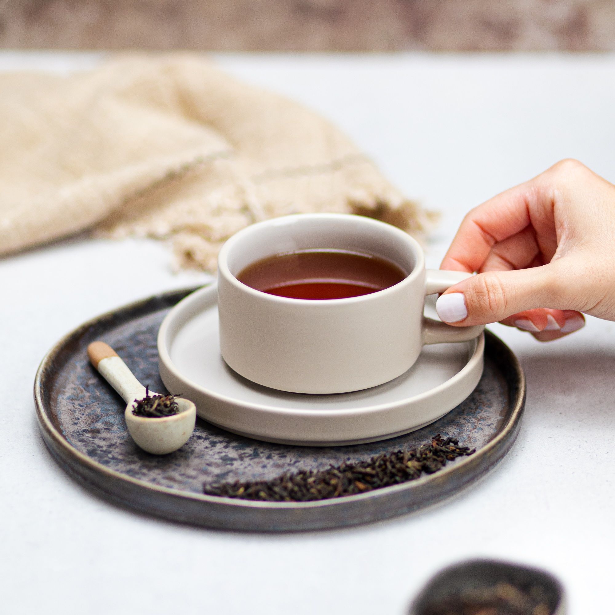 Tasse Tee auf Untertasse. Löffel mit Teeblättern. Hand greift nach Tasse. Dunkle Platte.