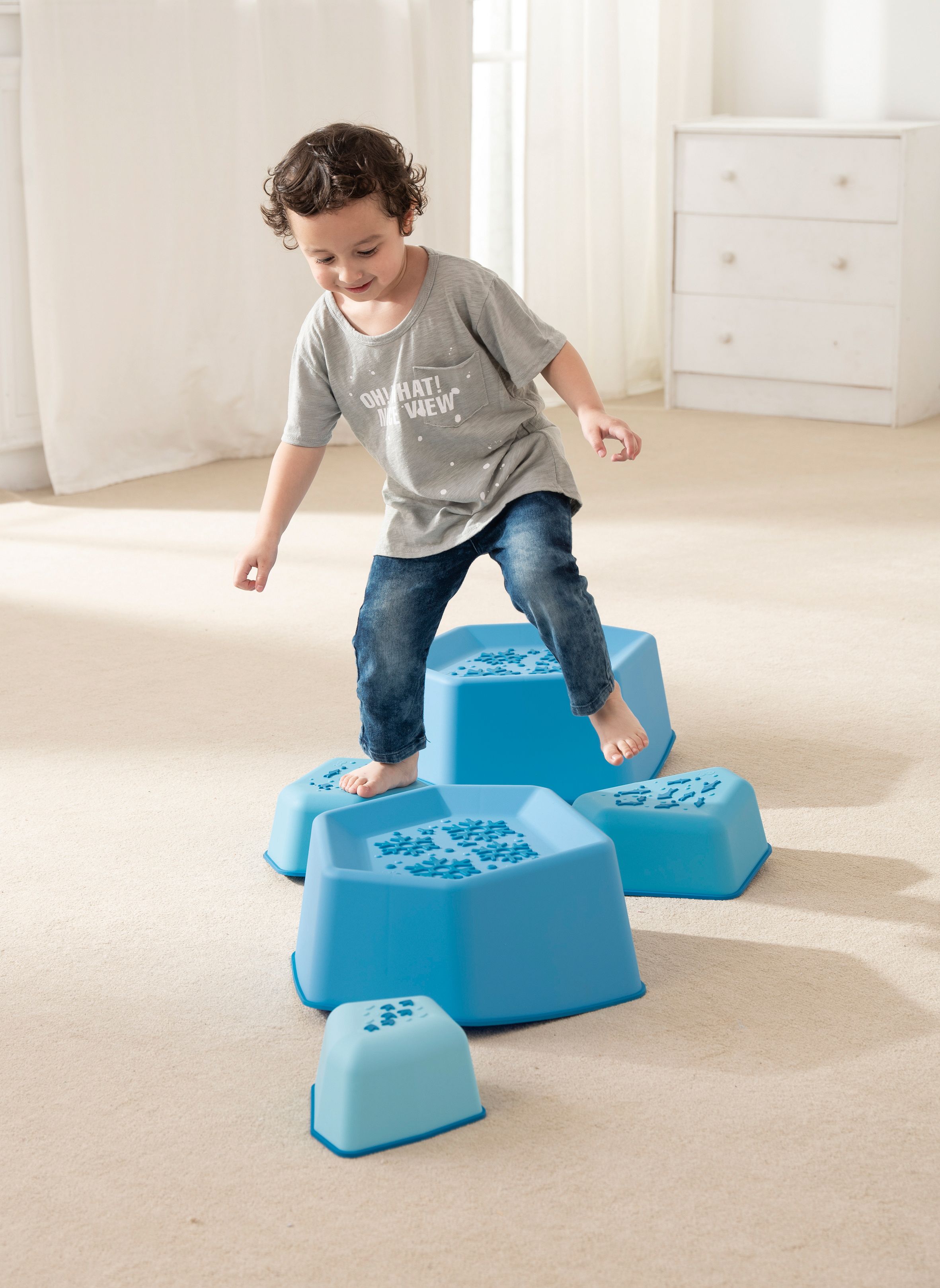 Kind balanciert auf blauen, sechseckigen Balance-Rockern mit Schneeflocken-Design. Auf Teppichboden.