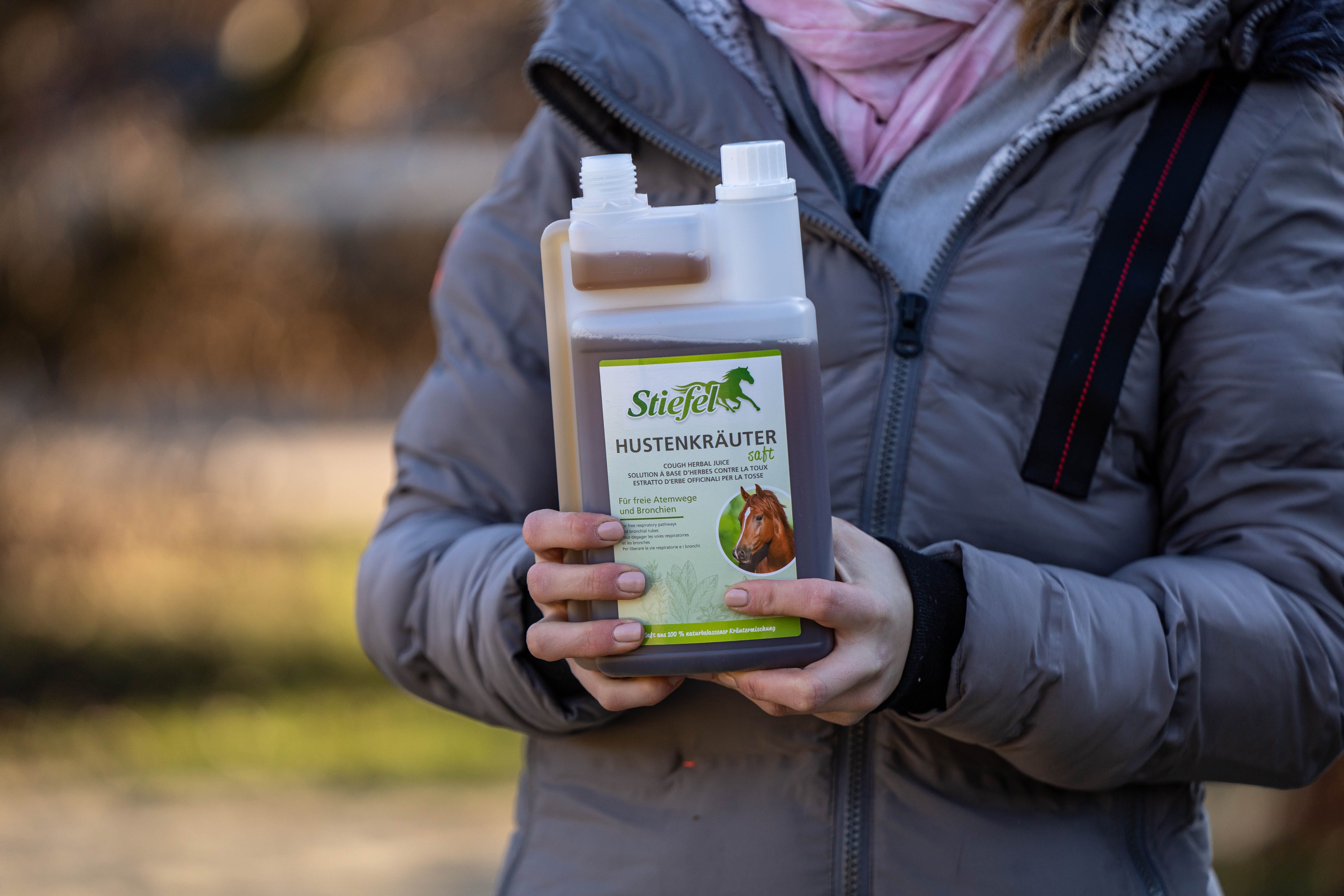 Person hält eine Flasche mit brauner Flüssigkeit. Auf dem Etikett steht der Produktname und ein Pferd. Die Flasche hat einen weißen Deckel.