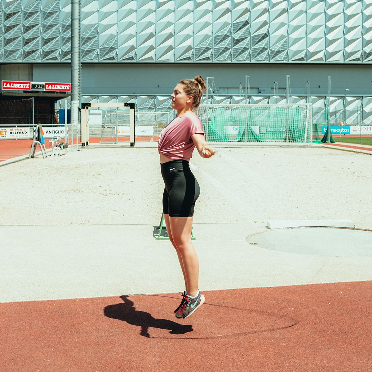 Frau springt mit einem Springseil auf einer Sportanlage. Rotes Feld, Betonboden, Gebäude im Hintergrund.
