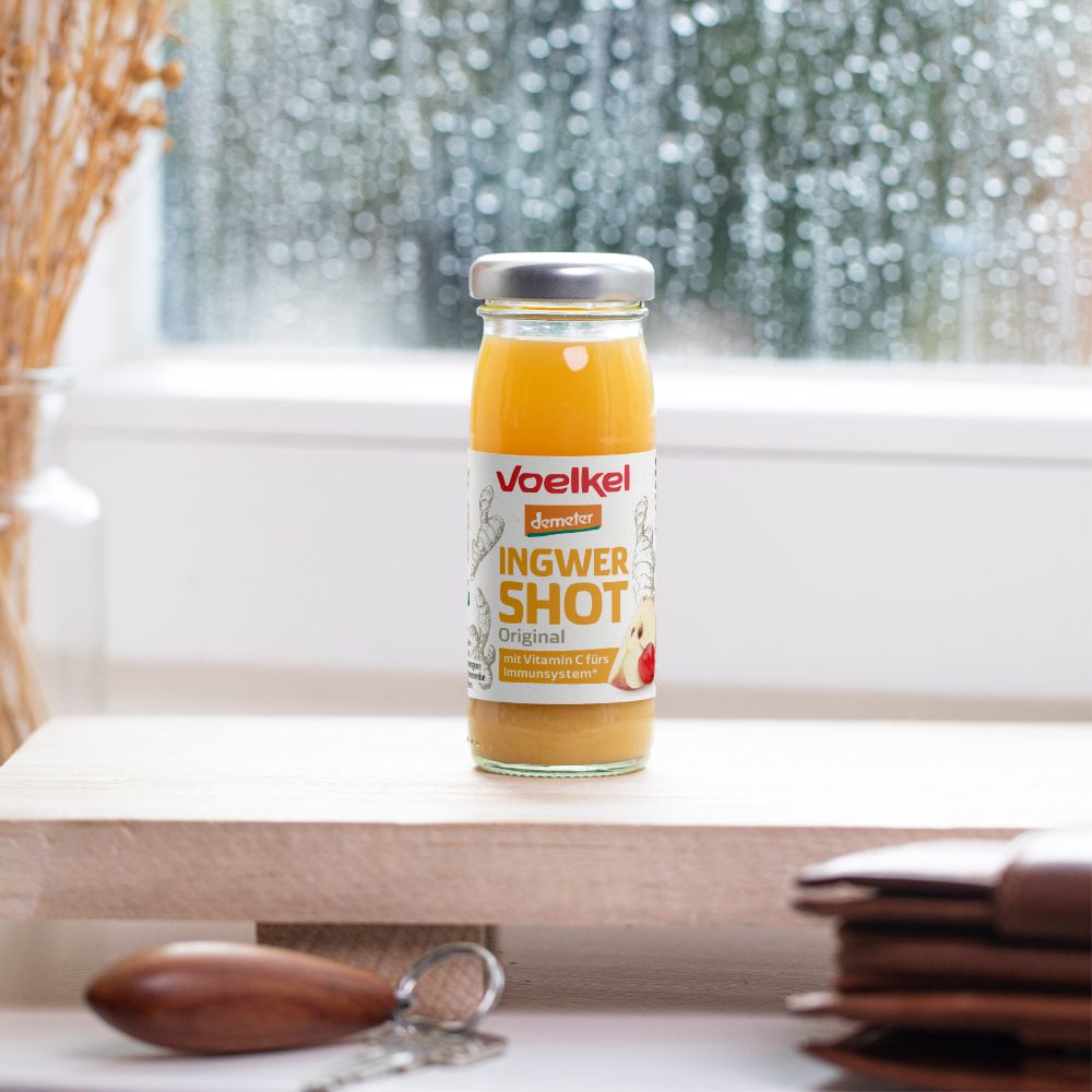 Glasflasche mit gelbem Saft, Etikett "Voelkel Ingwer Shot Original". Text: Vitamin C fürs Immunsystem. Auf Holzbrett, Fenster im Hintergrund.