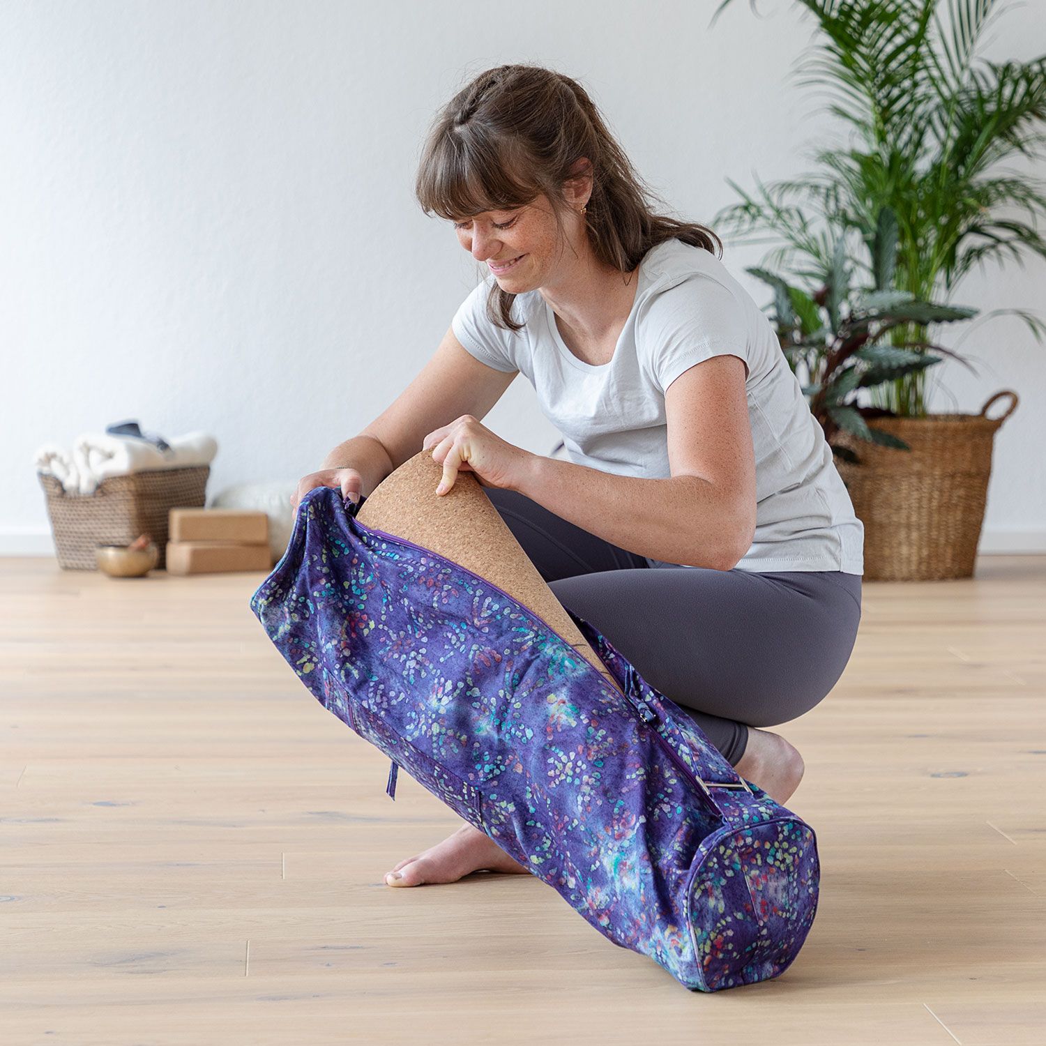 Femme ouvrant un sac de yoga. Sac lila à motifs colorés. Intérieur en liège. Sur sol en bois, dans une pièce claire.