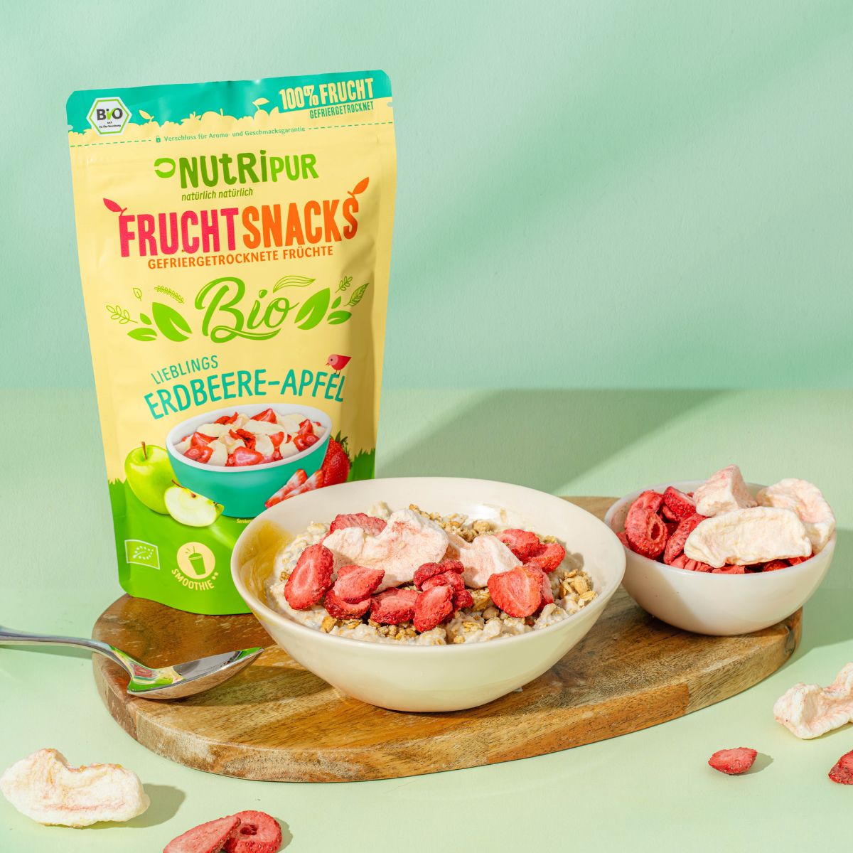 Beutel mit gefriergetrockneten Erdbeere-Apfel-Früchten. Schale mit Müsli und Früchten, daneben Schale mit Früchten. Bio-Siegel.