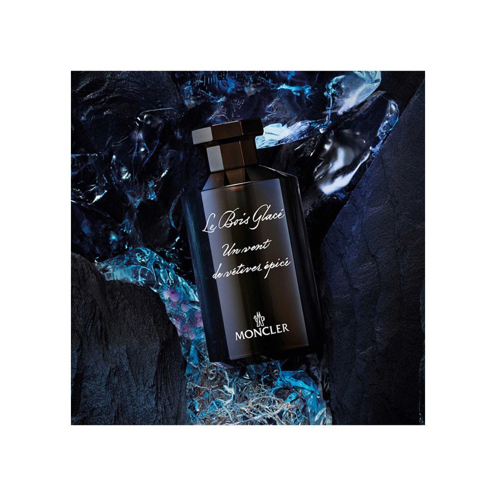 Dunkle Glasflasche mit schwarzem Verschluss. Aufschrift: Le Bois Glacé, Un vent de vétiver épicé, MONCLER. Auf blauem Untergrund.