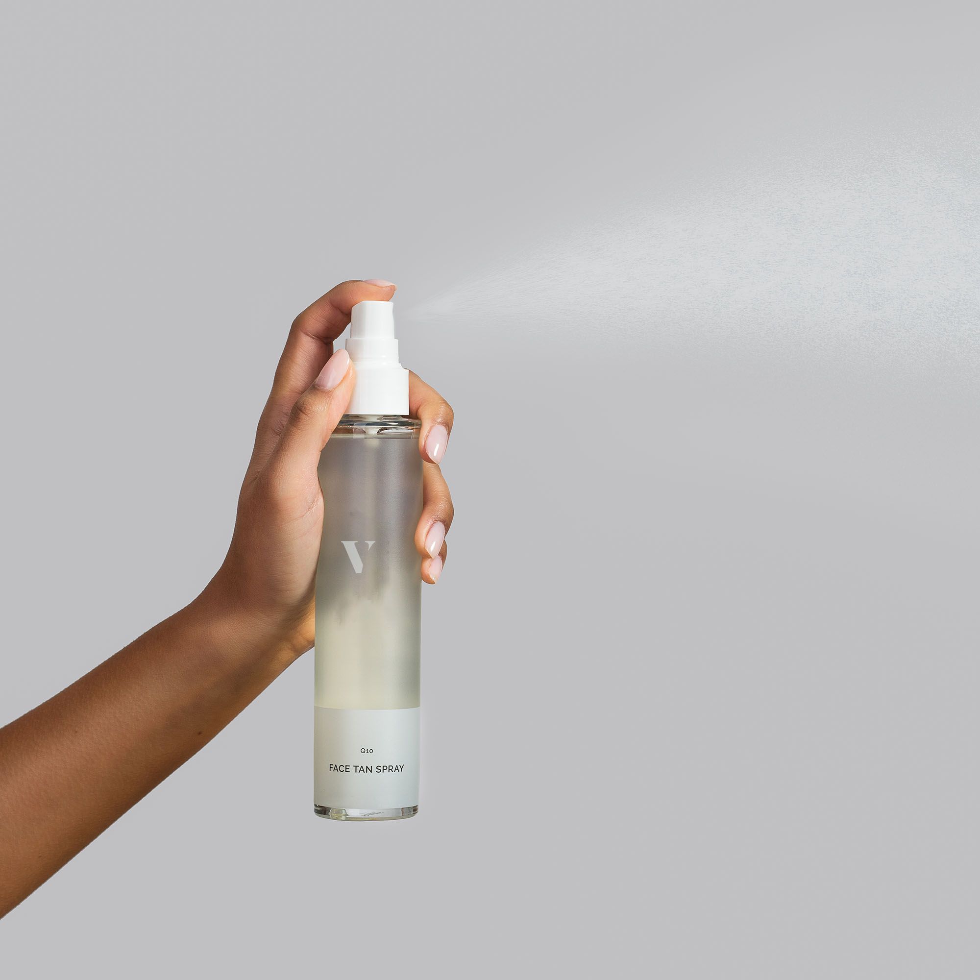 Hand sprüht transparente Flüssigkeit aus einer Sprühflasche. Aufschrift: FACE TAN SPRAY.