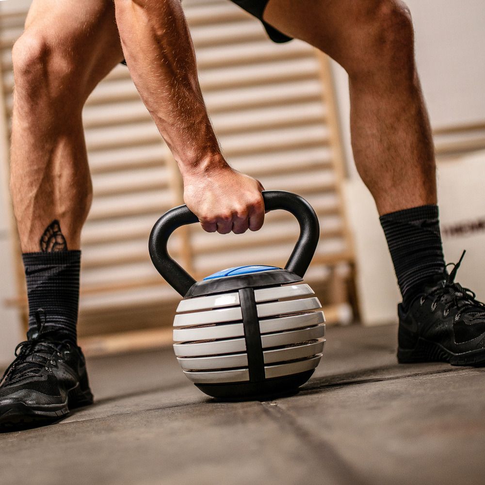 Kettlebell Vario, schwarz-weiß, Detailaufnahme. Griff und Scheiben sichtbar. Athlet hält das Gerät. Hintergrund: Fitnessraum.
