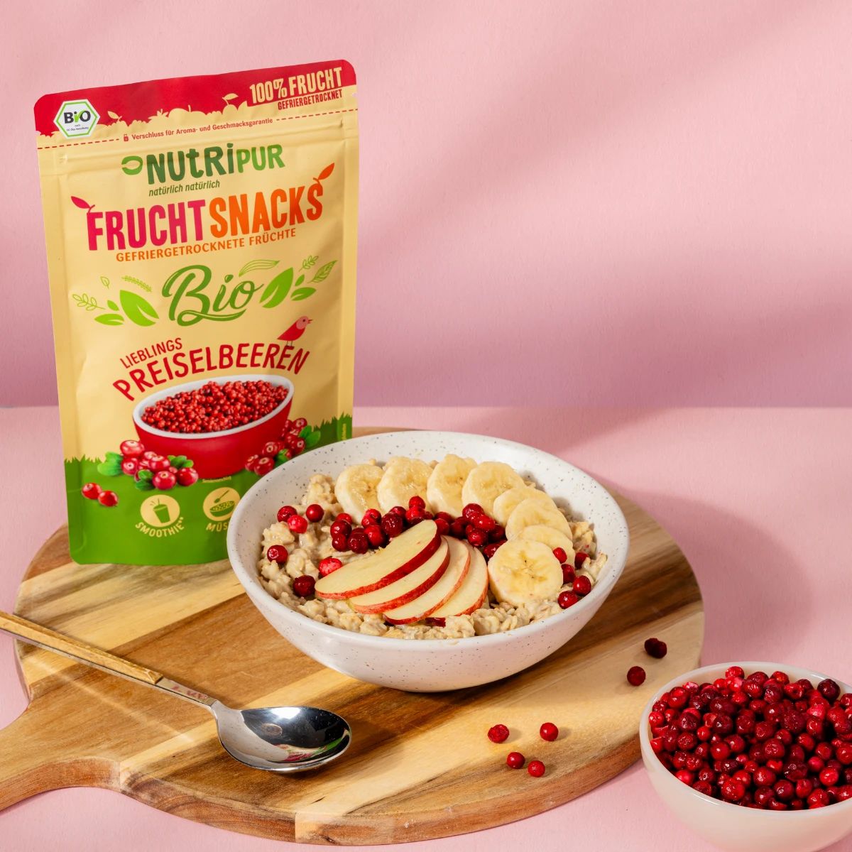 Schale mit Haferflocken, Bananen- und Apfelscheiben, Preiselbeeren. Daneben: Beutel mit Preiselbeeren, Schale mit Preiselbeeren.