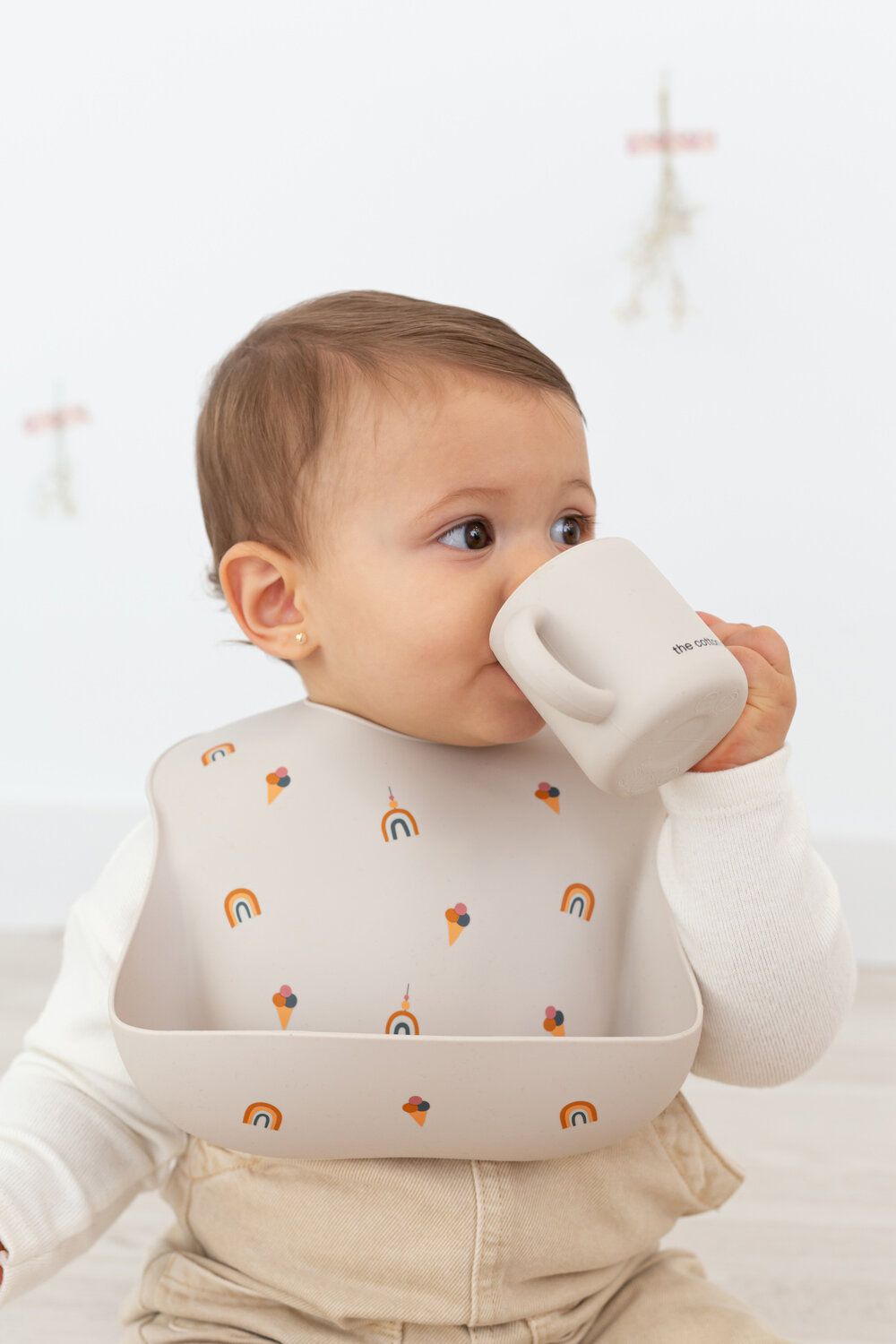 Baby trinkt aus einem hellen Silikonbecher mit Griffen. Baby trägt Lätzchen mit Eiscreme-Motiven.