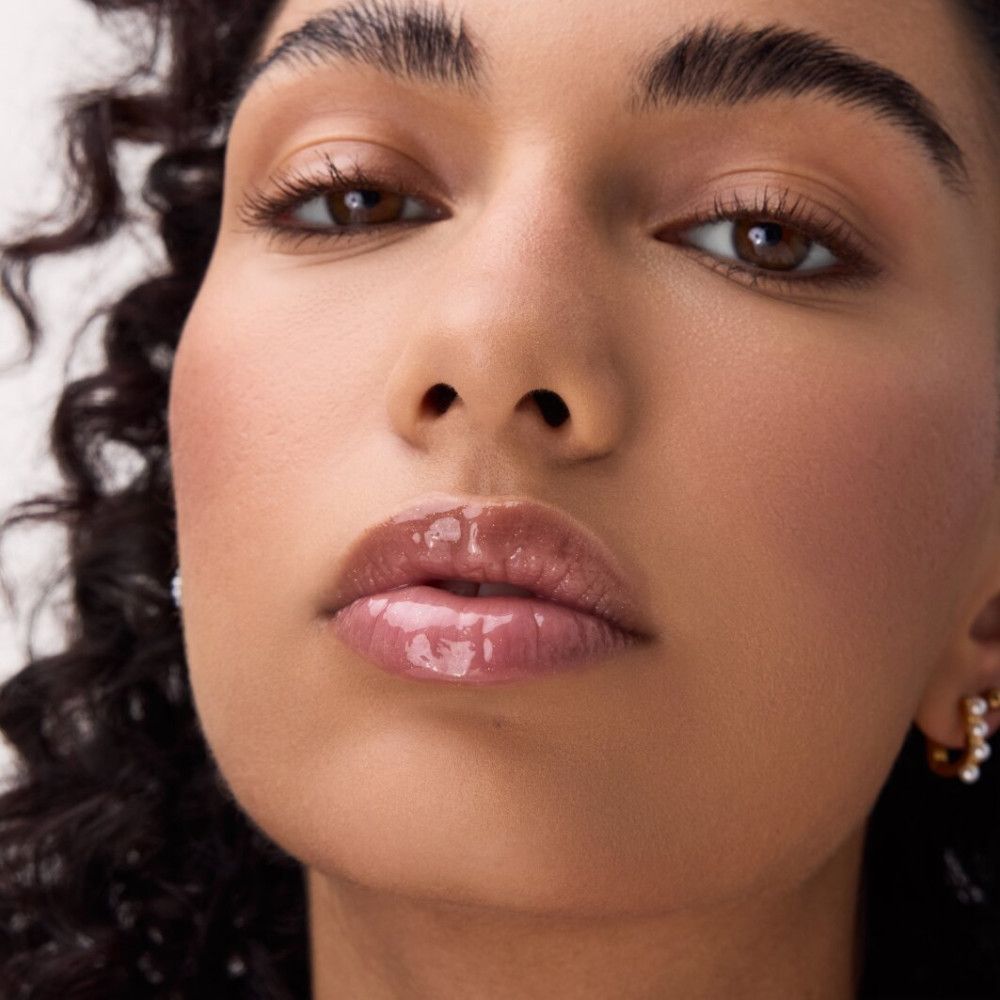 Nahaufnahme eines Gesichts mit glänzenden Lippen. Die Lippen haben einen rosafarbenen Farbton. Die Person hat lockiges Haar.