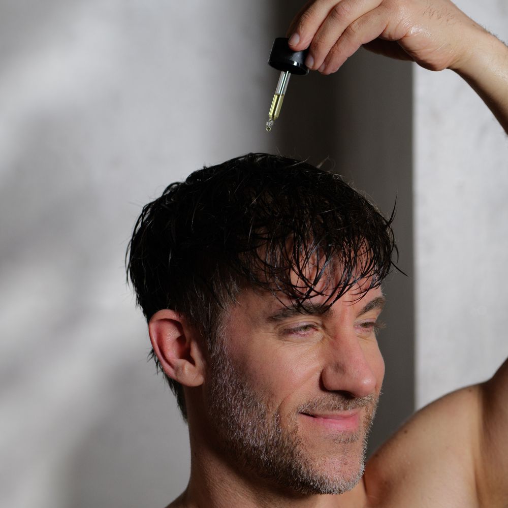 Mann mit Serum-Pipette. Serum wird auf Kopf getropft. Flasche mit schwarzem Deckel.